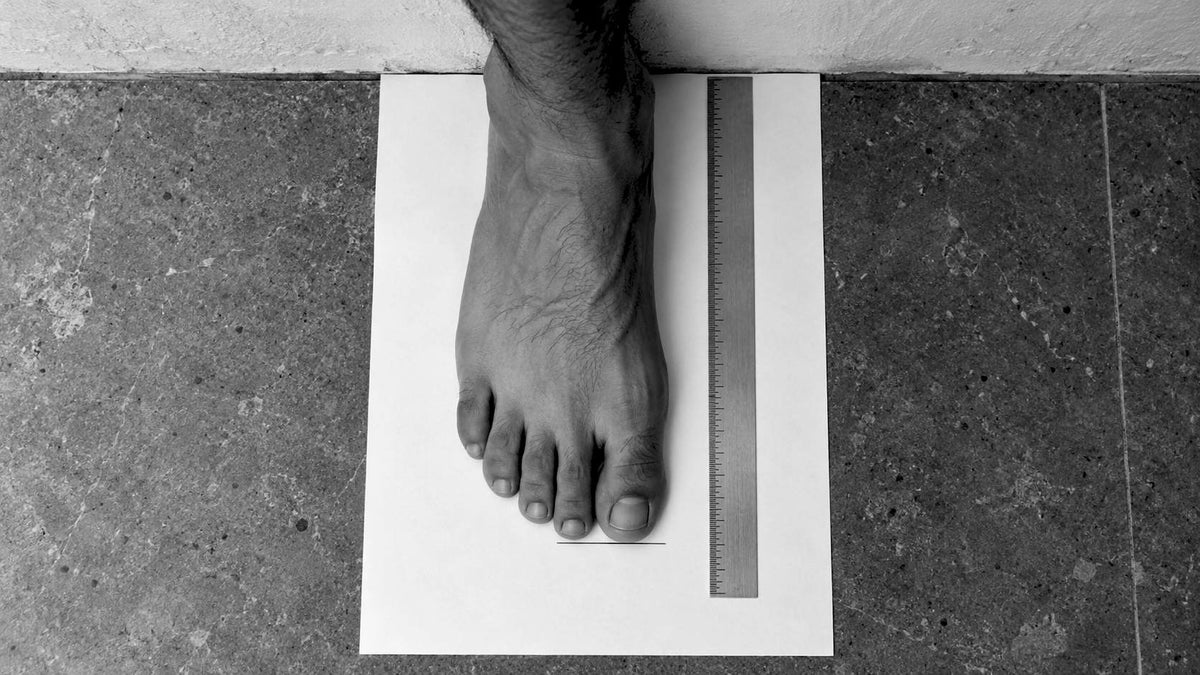 Black-and-white illustration, seen from above: a bare foot on a sheet of paper, heel to the wall, a pencil mark at the longest toe and a steel ruler alongside.