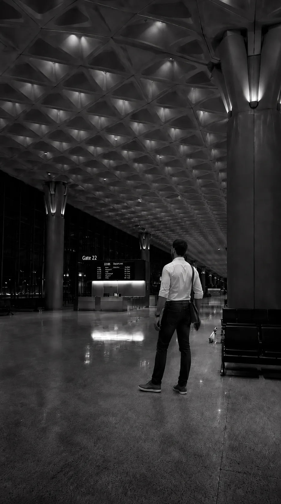 Indian airport departures hall, diamond-grid ceiling with column uplighting, single figure standing mid-frame in three-quarter back facing the departures board at 22:05