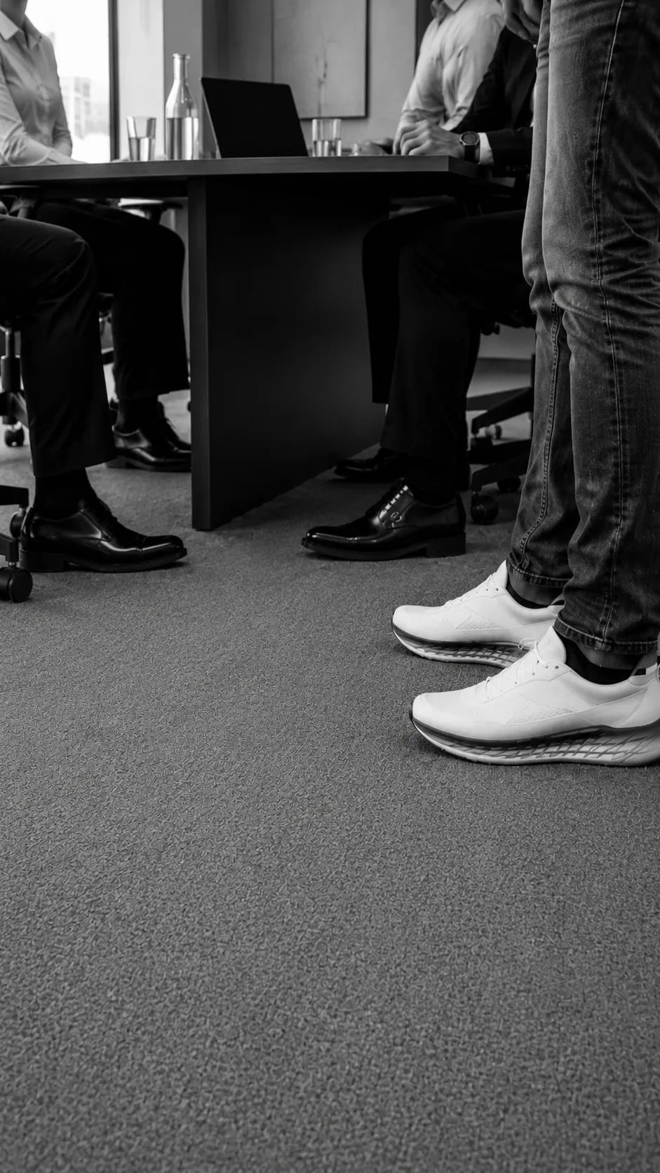 Three executives in formal shoes seated at a boardroom table, with a standing presenter visible knees-down in white performance sneakers.