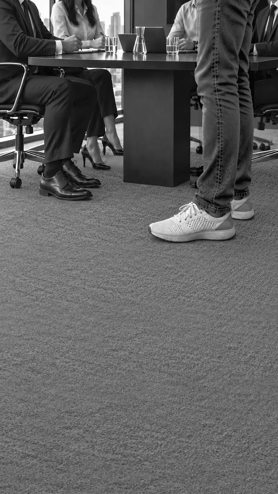 Four colleagues in formal shoes seated at a boardroom table, and a man standing beside them in jeans and white running shoes.