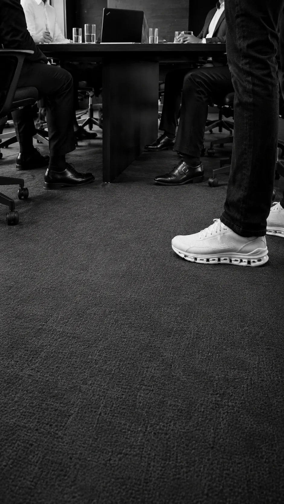Three executives in formal shoes seated at a boardroom table, with a standing presenter visible knees-down in white performance sneakers.